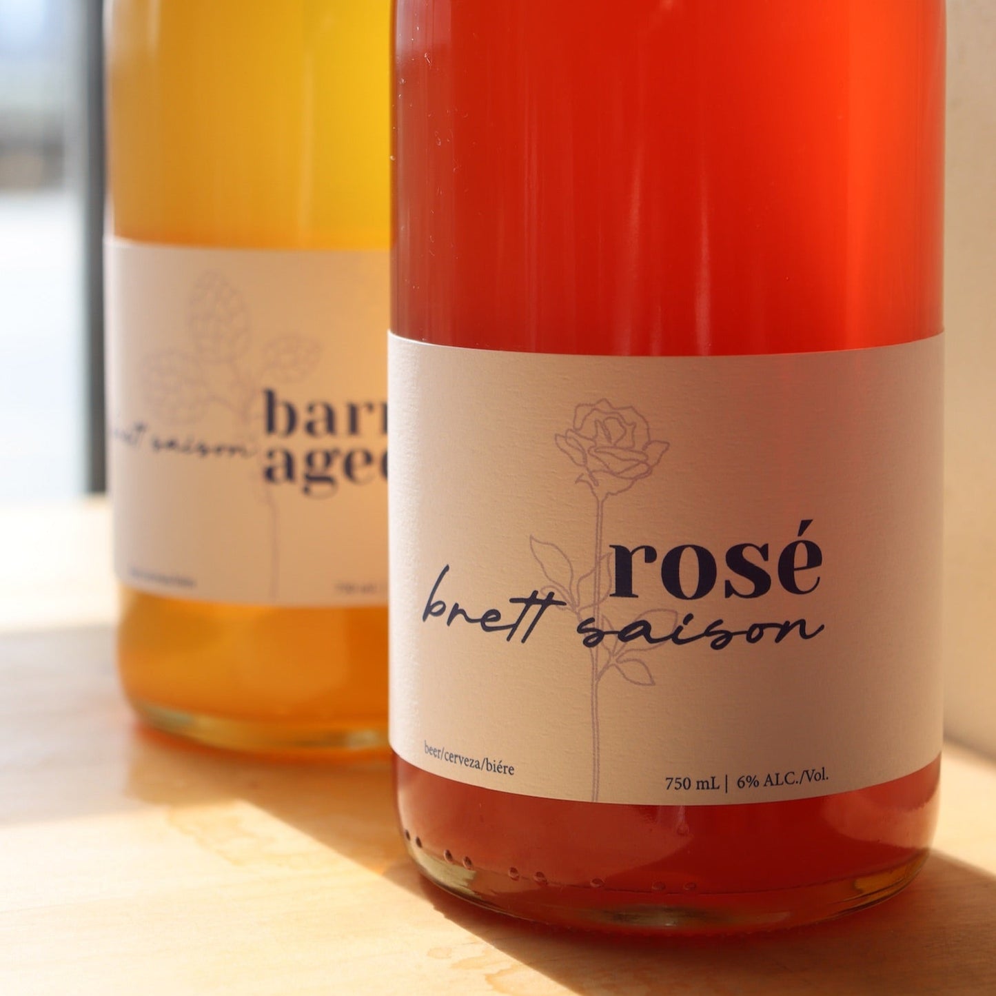 Two bottles of beer, one labeled 'Barrel Aged' and the other 'Rose Brett Saison', with branding and alcohol volume information visible.